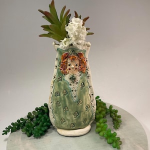 Puede incluir: Un jarrón de cerámica verde con una cara caprichosa y un patrón floral. El jarrón está decorado con detalles blancos y verdes y tiene una base pequeña y redonda. Está lleno de un pequeño arreglo de flores blancas y suculentas verdes.