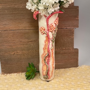 May include: A tall, cylindrical ceramic vase in an off-white glaze, featuring a vertical, textured orange and pink decorative strip with raised, sculpted orange flowers and green stems. The vase is filled with white flowers and stands upright.