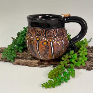 Könnte beinhalten: Eine handgefertigte Keramik-Tasse mit schwarzem Innenraum und Henkel. Die Außenseite weist ein braunes und orangefarbenes Muster mit weißen und gelben Akzenten auf. Die Tasse ist mit einer kleinen, hellfarbigen Verzierung am Henkel verziert.