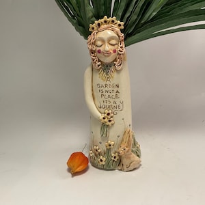 Puede incluir: Un jarrón de cerámica caprichoso con una cara serena, una corona y la inscripción "GARDEN IS NOT A PLACE ITS A JOURNEY". Presenta un conejo, flores y una linterna naranja. El follaje verde está en el jarrón.