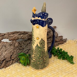 Puede incluir: Una jarra alta de cerámica pintada a mano con asa y borde azules. El cuerpo blanquecino presenta abetos verdes y estrellas amarillas. La tapa es azul con una parte superior texturizada verde y un adorno de estrella amarilla.