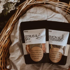 May include: Two black pouches containing simmer packs with white labels. One label reads Citrus & Bay, the other Lemon & Rosemary. Both feature a round tan sticker with instructions to pour contents into boiling water and reduce to a simmer.