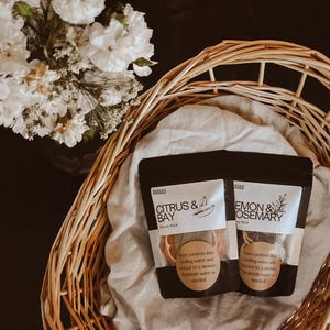 May include: Two black pouches labeled Citrus & Bay and Lemon & Rosemary containing dried fruit and herbs, placed inside a light brown woven wicker basket lined with a light gray cloth, next to a bouquet of white flowers on a dark surface.