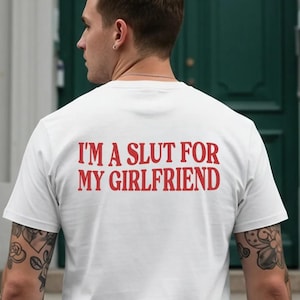 Może przedstawiać: Biała koszulka z czerwonym napisem "I'M A SLUT FOR MY GIRLFRIEND" wydrukowanym na plecach. Koszulka jest noszona przez osobę z tatuażami na ramieniu.