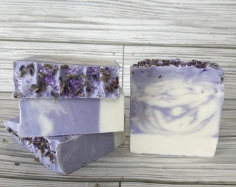 May include: Handmade lavender soap bars. The bars are a light purple color with white swirls and topped with dried lavender buds. The soap is stacked in a group of three bars and one bar is standing upright.