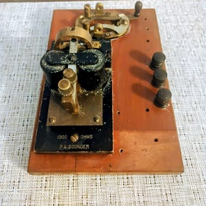 May include: A vintage telegraph sounder mounted on a rectangular wooden base. The device features black cylindrical coils, brass components, and three small knobs on the side. The black metal plate is labeled 1000 OHMS and 7 A SOUNDER.