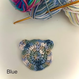 Puede incluir: Un aplique de oso azul tejido a ganchillo con hilos azules, verdes y beiges. Un ganchillo dorado y dos ovillos de lana colorida en el fondo. La palabra "Blue" está en la parte inferior.