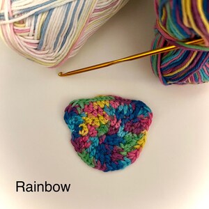 Może przedstawiać: Kolorowy projekt szydełkowy z tęczowym wzorem, złotym szydełkiem i dwiema kulkami przędzy. Słowo "Rainbow" jest napisane na dole obrazu. Przędza jest w odcieniach różu, niebieskiego, żółtego i zielonego.