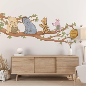 Op de afbeelding: Een Winnie de Poeh-thema wanddecoratie voor een kinderkamer met Winnie de Poeh, Biggetje, Teigetje en Eeyore-personages op een tak met een honingraat. De sticker is op een witte muur met een houten dressoir en een lamp.