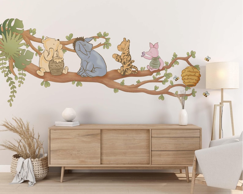 Op de afbeelding: Een Winnie de Poeh-thema wanddecoratie voor een kinderkamer met Winnie de Poeh, Biggetje, Teigetje en Eeyore-personages op een tak met een honingraat. De sticker is op een witte muur met een houten dressoir en een lamp.