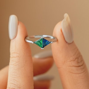 May include: A silver ring featuring a geometric, diamond-shaped gemstone with green and blue facets. The ring has a delicate band and small accent stones on either side of the central gem. The ring is held up by a person's fingers.