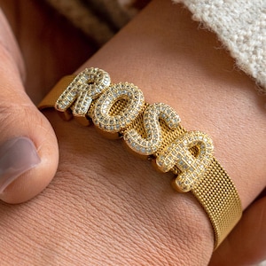 Op de afbeelding: Goudkleurige armband met de naam "ROSA" in sprankelende, diamantachtige letters. De armband heeft een mesh-ontwerp en de letters zijn bezet met kleine, heldere steentjes. De armband wordt om een pols gedragen, met een crème trui op de achtergrond.