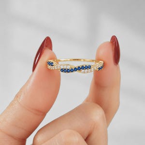 May include: A gold ring with a twisted design, featuring alternating rows of small blue and white gemstones. The ring is held between two fingers, showcasing its intricate details and craftsmanship.
