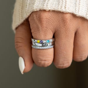 May include: Close-up of four silver rings stacked on a finger. The top three rings feature yellow, purple, and blue gemstones. The third ring is engraved with the word "NANA". The bottom ring is encrusted with small, clear stones.