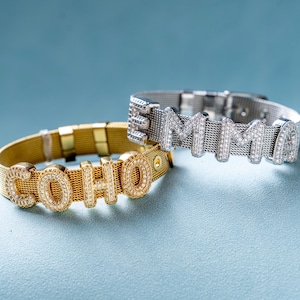 Könnte beinhalten: Zwei Armbänder aus Mesh, eines goldfarben und eines silberfarben, jeweils mit einem Namen, der in funkelnden, mit Edelsteinen besetzten Buchstaben geschrieben ist. Das goldene Armband schreibt "SOHO", das silberne Armband "EMMA".