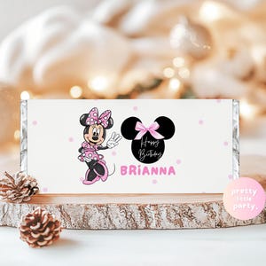 Puede incluir: Envoltorio blanco de barra de caramelo con Minnie Mouse en rosa y negro. El envoltorio tiene el texto "Happy Birthday Brianna" y lunares rosas. Se muestra sobre una rodaja de madera.