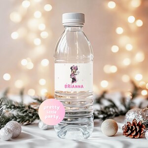 Puede incluir: Una botella de agua de pl&aacute;stico transparente con una tapa blanca y una etiqueta con un dise&ntilde;o de dibujos animados de Minnie Mouse. La etiqueta incluye el nombre "Brianna" en texto rosa. El fondo tiene luces bokeh y decoraciones festivas.