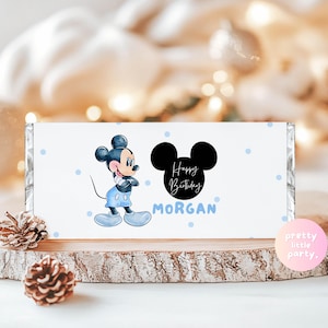 Puede incluir: Envoltorio blanco de barra de caramelo con Mickey Mouse con ropa azul, la frase "Happy Birthday MORGAN" y una silueta negra de Mickey Mouse. Lunares azul claro decoran el envoltorio. Presentado sobre una rodaja de madera.