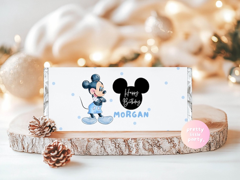 Puede incluir: Envoltorio blanco de barra de caramelo con Mickey Mouse con ropa azul, la frase "Happy Birthday MORGAN" y una silueta negra de Mickey Mouse. Lunares azul claro decoran el envoltorio. Presentado sobre una rodaja de madera.