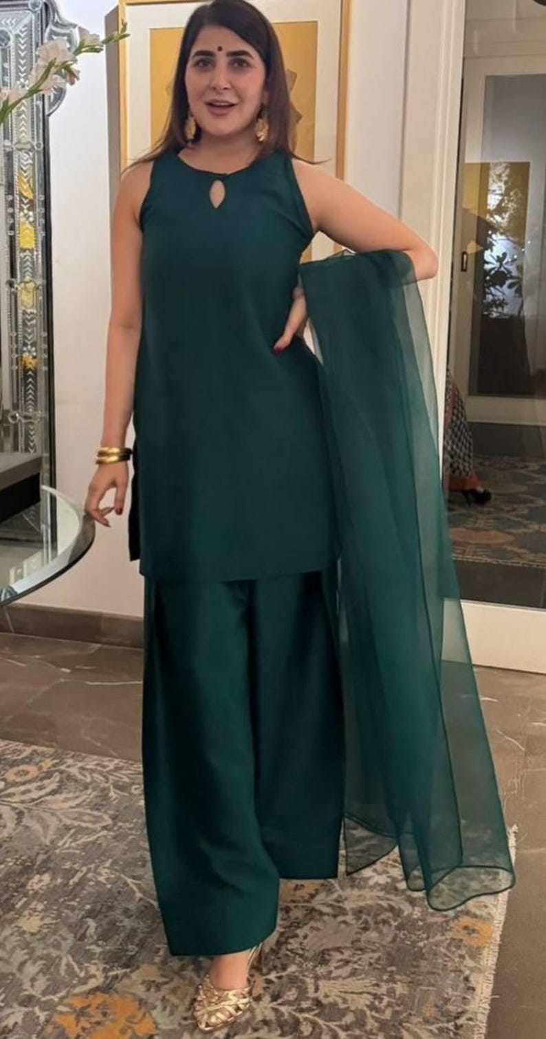 May include: A woman wearing a dark green sleeveless kurta with a matching palazzo pant and a sheer dupatta. She is wearing gold bangles and gold earrings. The outfit is a traditional Indian style.