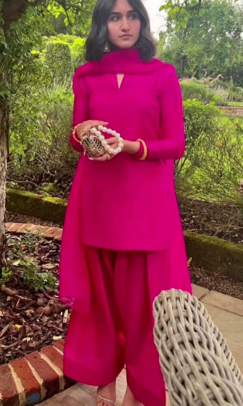 May include: A woman wearing a bright pink long-sleeved top and matching trousers. The top has a high neck with a small keyhole opening. She is holding a gold and pearl clutch.