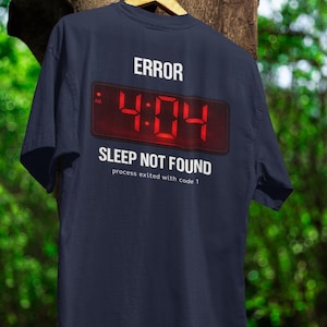 May include: Navy blue t-shirt featuring the word "ERROR" above a digital clock display showing "4:04" in red. Below, it reads "SLEEP NOT FOUND" and "process exited with code 1". The shirt is hanging on a wooden hanger.
