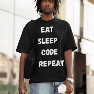 Puede incluir: Camiseta negra con las palabras "EAT SLEEP CODE REPEAT" en letras blancas. La persona lleva vaqueros azul claro y un reloj plateado. También se ven un bolso negro y una taza de café blanca.