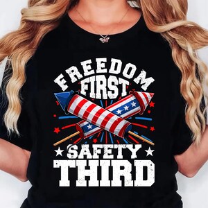 May include: Black t-shirt with a graphic design featuring two crossed firework rockets in red, white, and blue. The text reads "FREEDOM FIRST" above and "SAFETY THIRD" below the rockets, with star accents.