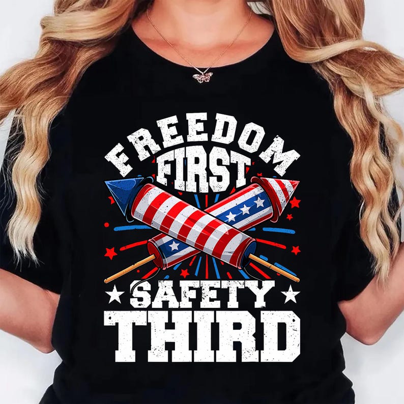 May include: Black t-shirt with a graphic design featuring two crossed firework rockets in red, white, and blue. The text reads "FREEDOM FIRST" above and "SAFETY THIRD" below the rockets, with star accents.