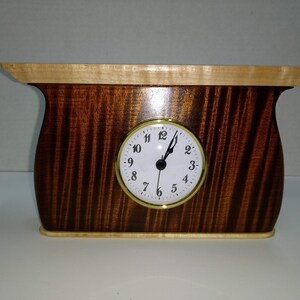 May include: A handcrafted wooden clock with a light-colored top and base, and a dark, patterned body. The clock face is white with black numerals and hands, set in a gold-colored frame. The clock is approximately 10 inches wide.