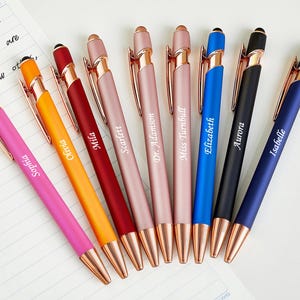 May include: An assortment of personalized pens in various colors, including pink, orange, red, and blue. Each pen has a metallic rose gold accent and is engraved with a name. The pens are arranged on a white surface.
