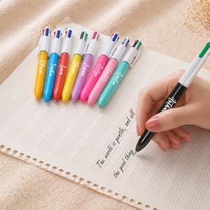 May include: A collection of colorful multi-color pens with names printed on them, arranged on a sheet of paper. One pen is held by a hand, writing the words "The world is gentle, and all the good thing."
