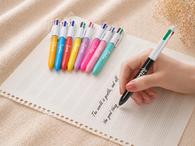 May include: A collection of colorful multi-color pens with names printed on them, arranged on a sheet of paper. One pen is held by a hand, writing the words "The world is gentle, and all the good thing."