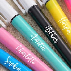 May include: A close-up of several colorful markers with white tips and the names "Sophia", "Charlotte", "Amelia", "Arthur", "Harper", and "Evelyn" in white script. The markers are in shades of blue, pink, teal, black, yellow, and pink.