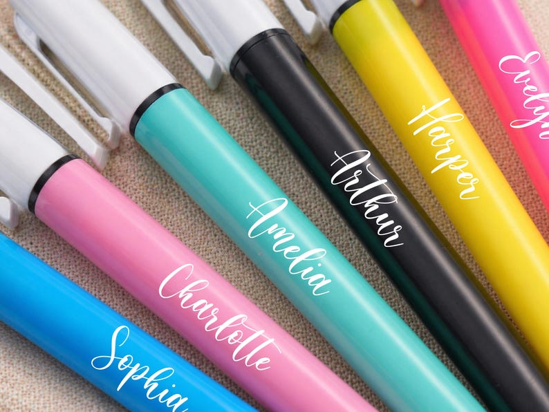 May include: A close-up of several colorful markers with white tips and the names "Sophia", "Charlotte", "Amelia", "Arthur", "Harper", and "Evelyn" in white script. The markers are in shades of blue, pink, teal, black, yellow, and pink.