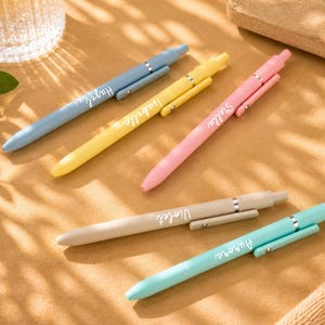 May include: An assortment of colorful pens in shades of blue, yellow, pink, beige, and teal. Each pen has a silver clip and a name printed on the side: Hazel, Isabella, Stella, Violet, and Aurora. The pens are arranged on a brown surface.