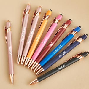 May include: A collection of personalized pens in various colors, including pink, orange, red, blue, and black. Each pen has a gold-colored clip and tip, with names like "Scarlett" and "Aurora" engraved on them.