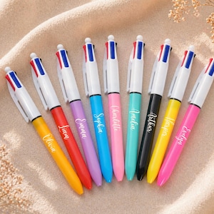 Op de afbeelding: Een assortiment kleurrijke balpennen met witte barrels en namen op de zijkanten. Elke pen heeft een andere kleur inkt en een bijpassende gekleurde barrel. De pennen liggen op een beige ondergrond.