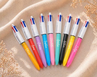 May include: A collection of multi-coloured ballpoint pens with white barrels and names printed on the sides. Each pen has a different ink colour and a matching barrel. The pens are displayed on a beige surface.