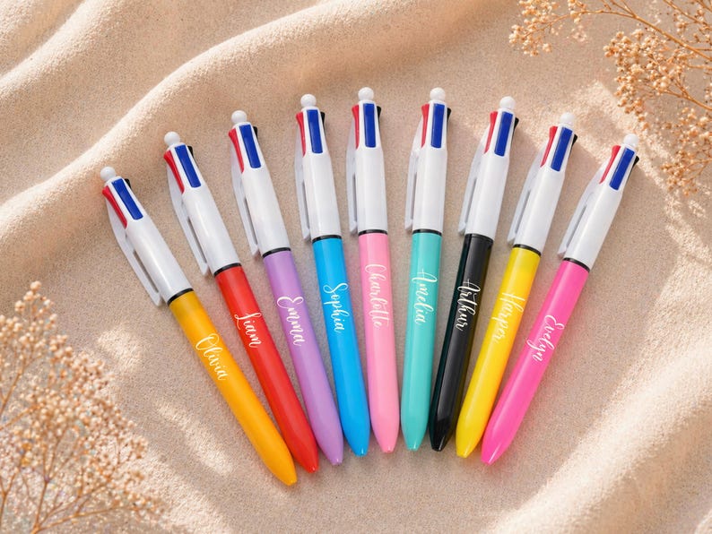 May include: An assortment of colorful ballpoint pens with white barrels and names printed on the sides. Each pen has a different colored ink and a matching colored barrel. The pens are arranged on a beige surface.