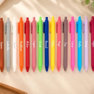 Könnte beinhalten: Eine Sammlung farbenfroher Kugelschreiber, jeder mit einem anderen Namen auf der Seite. Die Stifte sind in verschiedenen Farben erhältlich, darunter Türkis, Braun, Grau, Weiß, Orange, Rot, Rosa, Grün, Blau, Gelb und Schwarz. Die Namen sind kursiv geschrieben.