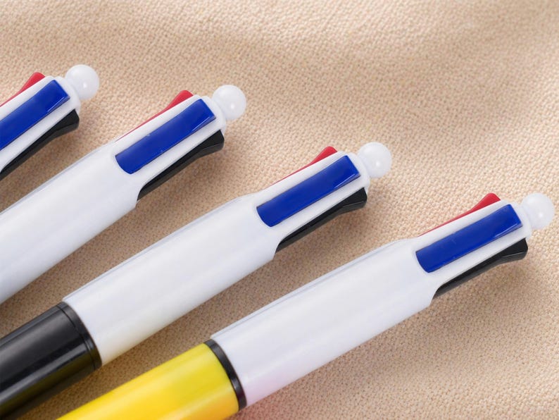 May include: Close-up of several multi-color pens. Each pen has a white barrel with a different colored button on top. The pens have black, red, blue, and yellow accents. The pens are arranged diagonally on a beige surface.