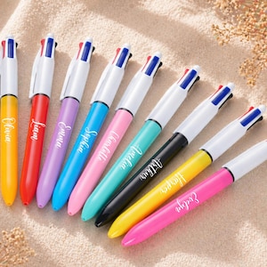 May include: A collection of colorful ballpoint pens with white barrels and four-color ink selection buttons. Each pen is a different color and has a name printed on the side. The pens are arranged diagonally on a light-colored surface.