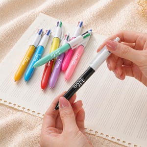 May include: A collection of colorful multi-color pens with white barrels and black tips, each with a different color ink. The pens are arranged on a lined paper, with one pen held by a person. The pens are yellow, blue, green, red, purple, and pink.