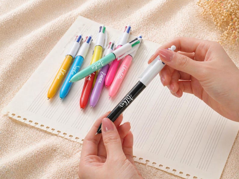 May include: A collection of colorful multi-color pens with white barrels and black tips, each with a different color ink. The pens are arranged on a lined paper, with one pen held by a person. The pens are yellow, blue, green, red, purple, and pink.