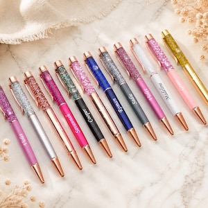 May include: A collection of personalized pens with glitter-filled barrels in various colors, including pink, silver, and blue. Each pen has a rose gold tip and clip, with names like "Violet" and "Madeline" printed on the side. The pens are arranged on a marble surface.
