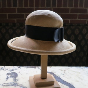 May include: A beige straw hat with a black ribbon and bow. The hat is displayed on a wooden stand.