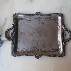May include: A silver tray with ornate handles and a slightly worn surface. The tray has a rectangular shape with rounded corners and a decorative border.