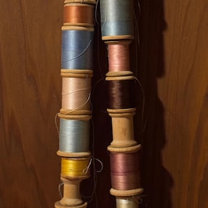 Puede incluir: Una colección de carretes de hilo, enrollados en bobinas de madera, en varios colores, incluyendo rosa, marrón, azul y blanco. Los carretes están dispuestos en dos filas verticales.