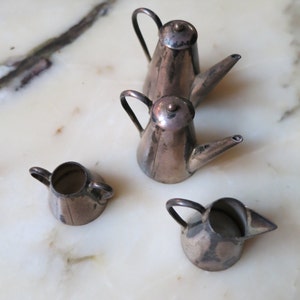 May include: Four silver miniature teapots with handles and spouts. The teapots are arranged on a white marble surface.